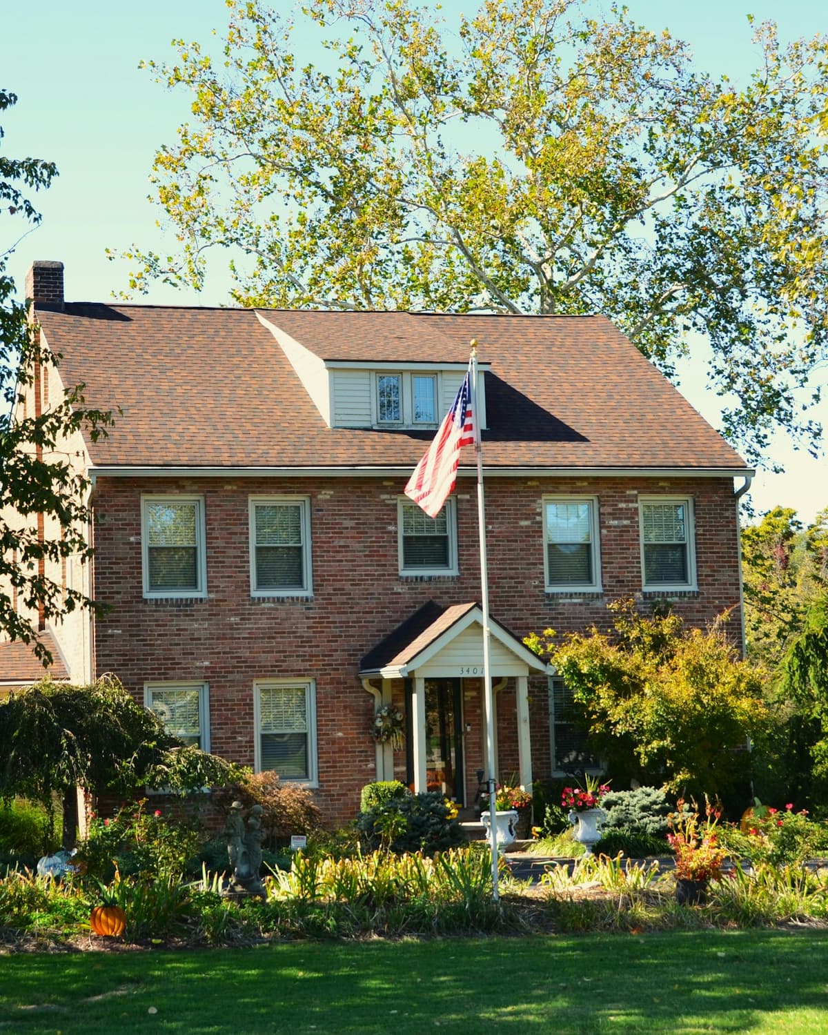 Red brick colonial house with an American flag, surrounded by autumn trees and landscaped garden beds